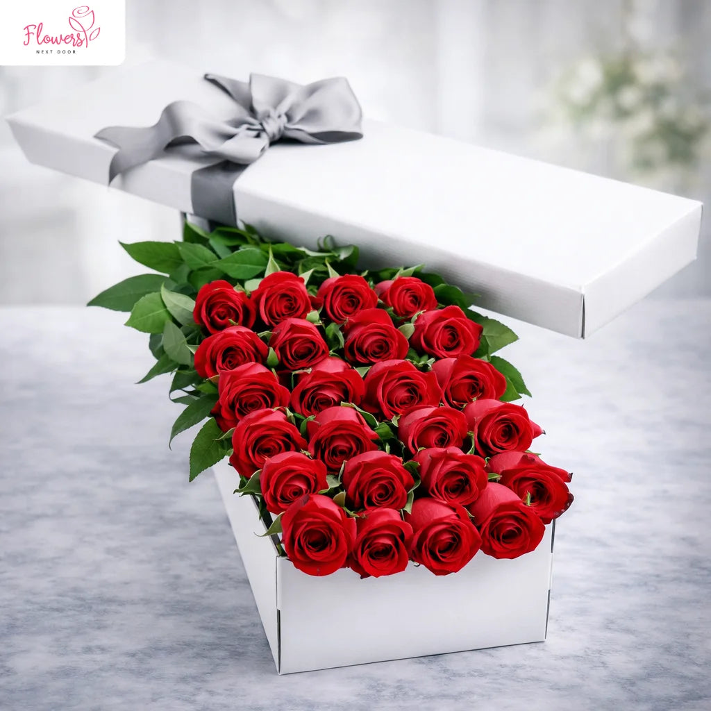 Heart-shaped arrangement of red roses in a white box with a gray ribbon on a light gray background.