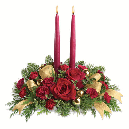Decorative Christmas arrangement with red candles, red roses, gold ribbons, and greenery on a white background.